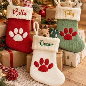 May include: Three knit Christmas stockings with white cuffs. The red stocking features the name Bella and a white paw print. The green stocking features the name Toby and a red paw print. The white stocking features the name Oscar and a red paw print.