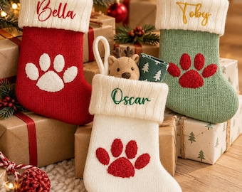 Puede incluir: Tres calcetines navideños de punto con puños blancos. El calcetín rojo lleva el nombre Bella y una huella blanca. El verde lleva el nombre Toby y una huella roja. El blanco lleva el nombre Oscar y una huella roja.