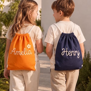 May include: Two drawstring backpacks, one bright orange with the name Amelia and a small flower appliqué, and one navy blue with the name Henry embroidered in white script. Both bags feature white cord straps and are worn on the back.