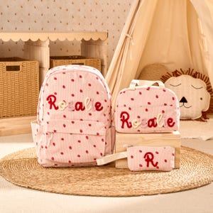 May include: A pink corduroy backpack, lunch bag, and pencil case set featuring a repeating strawberry pattern. Each item is personalized with the name Rosalie in red and light pink embroidered lettering, with the pencil case showing the letter R.