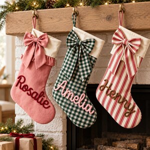 Pode incluir: Três meias de Natal penduradas numa lareira de madeira. A esquerda é rosa sólida com o nome Rosalie, a central é xadrez verde e branco com o nome Amelia, e a direita é listrada de vermelho e branco com o nome Henry.