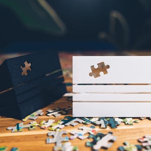 May include: A black, angled puzzle box and a white, rectangular puzzle box with a puzzle piece cutout. The boxes sit on a wooden surface scattered with puzzle pieces. The puzzle boxes are designed to hold puzzle pieces.