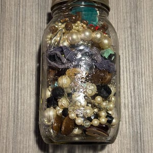 Op de afbeelding: Een glazen pot gevuld met een verzameling kralen, parels en stukjes stof. De pot heeft een metalen deksel en bevat een mix van kleuren en texturen, waaronder witte, zwarte, bruine en iriserende kralen. De inhoud is dicht opeengepakt in de pot.