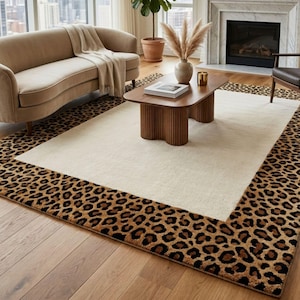 May include: A rectangular area rug featuring a large, solid cream-coloured centre panel surrounded by a wide border with a brown and black leopard print pattern. The rug is made of a soft, textured material and covers a portion of the wooden floor.