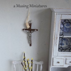May include: A small wooden crucifix with a silver Jesus figure hanging on a white wall. A white wicker chair with yellow flowers is in the foreground. The text "a Musing Miniatures" is at the top of the image.