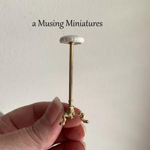 May include: Miniature gold-colored display stand with a round, white top. The stand has an ornate base and a slender stem. The text "a Musing miniatures" is above the stand.