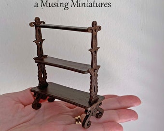 May include: A miniature brown wooden shelf with three tiers, held in a hand. The shelf has ornate, decorative supports and a scrolled base. The text "a Musing Miniatures" is in the upper left corner.