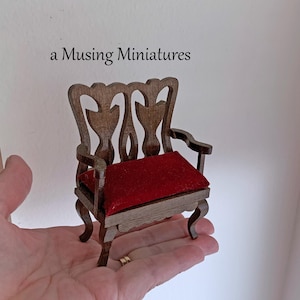 May include: Miniature wooden chair with a red velvet seat. The chair has an ornate back and curved legs. The text "a Musing Miniatures" is above the chair.