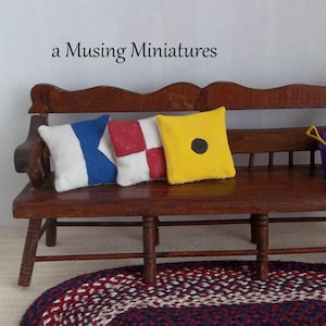 May include: Miniature nautical-themed pillows in white, blue, red, and yellow rest on a wooden bench. The pillows feature signal flag designs and a black dot. The bench sits on a red, white, and blue braided rug. Text on the wall reads "a Musing Miniatures."