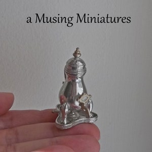 May include: A miniature silver-colored coffee pot with a gold-colored top and accents. The coffee pot sits on a small tray with a handle. The text "a Musing Miniatures" is above the coffee pot.