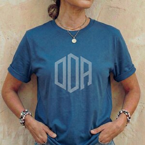 May include: A blue t-shirt with rolled-up sleeves featuring a light gray geometric design. The shirt is a solid color and has a crew neck. The model is wearing a gold necklace and bracelets.