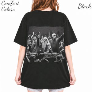 May include: A black t-shirt featuring a graphic print on the back. The print shows a black and white scene of a bearded figure and a bald figure behind a DJ deck with a laptop. The text Comfort Colors and Black appears in the corners.