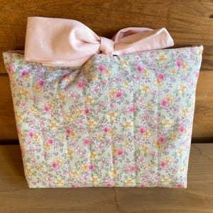 May include: A quilted fabric pouch featuring a light green background with a repeating floral pattern of small pink and yellow flowers. The top of the pouch is adorned with a large, solid light pink fabric bow attached to the center.
