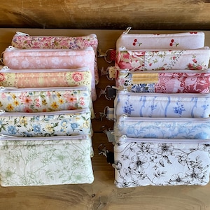 May include: A collection of rectangular fabric pouches with zippers and key rings. The pouches feature various floral patterns in colors like pink, yellow, blue, and green. Some have patchwork designs, while others display cherry or black and white floral prints.