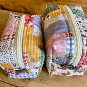 May include: Two patchwork fabric zippered pouches. The pouches are made of various floral and patterned fabrics in shades of pink, blue, yellow, and green. The zippers are cream-coloured. The pouches are sitting on a wooden surface.