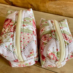 May include: Two quilted fabric pouches with floral patterns and cream-colored zippers. The pouches feature a patchwork design with red roses, green stripes, and cream-colored backgrounds. The pouches are likely used for storing small items.