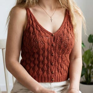 May include: A rust-colored, knitted camisole with a V-neck and button-up front. The top features a textured, openwork design and is made of a soft, breathable material. The camisole is sleeveless and has a fitted silhouette, perfect for layering or wearing on its own.