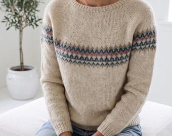 May include: A beige speckled knit jumper featuring a round neckline and long sleeves. The chest area displays a horizontal band with a geometric pattern in shades of muted blue, dusty pink, and dark brown, set against the light oatmeal-coloured yarn.