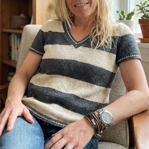 May include: A short-sleeved, striped V-neck top in grey and beige. The top has horizontal stripes and a ribbed trim. The person is wearing a watch and bracelets. The top is made of a knitted material.