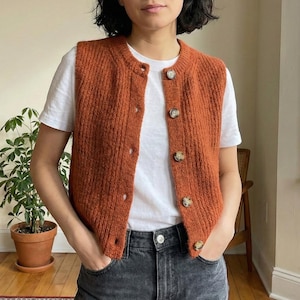 Peut inclure: Gilet tricoté de couleur rouille avec une fermeture boutonnée sur le devant. Le pull sans manches a une texture côtelée et une encolure ronde. Les boutons sont de couleur marron clair. Le gilet est porté sur un t-shirt blanc et associé à un jean en denim noir.