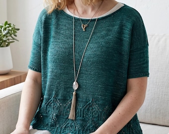 May include: A short-sleeved, dark teal knitted top featuring a wide, intricate lace-patterned hem. The garment is layered over a white base and is accessorised with a long, thin gold-toned chain necklace featuring a pale stone and a tassel pendant.