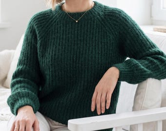 May include: A dark forest green long-sleeved jumper featuring a chunky ribbed knit texture, a round neckline, and raglan sleeves. The jumper is paired with white trousers and a delicate gold chain necklace with a small circular pendant.