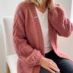 May include: A dusty rose open-front cardigan featuring a chunky knit texture, long sleeves with vertical cable knit detailing, and a ribbed trim along the front opening. A small rectangular white label with black text is attached to the inner collar.