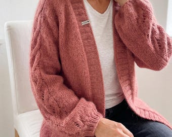 May include: A dusty rose open-front cardigan featuring a chunky knit texture, long sleeves with vertical cable knit detailing, and a ribbed trim along the front opening. A small rectangular white label with black text is attached to the inner collar.