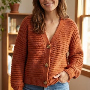May include: An orange-brown, ribbed-knit cardigan with a V-neck and three large, round, brown buttons. The long-sleeved sweater has a relaxed fit and is made of a soft, textured material. The cardigan is a cozy, everyday garment.