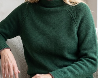 May include: A dark forest green long-sleeve jumper featuring a mock neck collar, raglan sleeves with visible knit detailing along the shoulder seams, and thin, contrasting maroon trim along the neckline, sleeve cuffs, and bottom hemline.