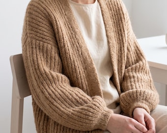 May include: A tan, open-front cardigan featuring a chunky ribbed knit texture, long sleeves with gathered cuffs, and a relaxed fit. The garment is worn over a plain, light-coloured top, showcasing the vertical knit pattern and soft, neutral tone.