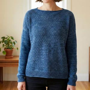 Puede incluir: Un suéter azul tejido con un patrón texturizado. El suéter tiene cuello barco y mangas largas. Las mangas y el dobladillo inferior tienen un diseño acanalado. El suéter se usa con pantalones negros. El fondo incluye una mesa de madera y una planta en maceta.