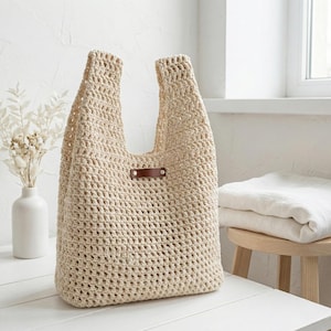 Puede incluir: Un bolso tote de crochet beige de tejido abierto con dos asas. Una pequeña etiqueta de cuero marrón está adherida al frente. La bolsa está sobre una superficie blanca. Un jarrón blanco con flores secas está a la izquierda.
