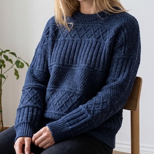 May include: A long-sleeved navy blue sweater featuring a crew neckline and a complex knit pattern. The design incorporates alternating horizontal bands of diamond-shaped cables, vertical ribbing, and a textured seed stitch throughout the garment.