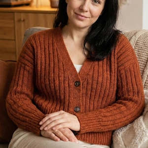 May include: A rust-colored, ribbed knit cardigan with a V-neck and three dark brown buttons. The long-sleeved sweater is made of a soft, textured material. The cardigan is a cozy and stylish piece of clothing.