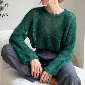 Puede incluir: Un suéter de punto suelto de color verde oscuro, de manga larga, con textura difusa y cuello redondo. El suéter es semitransparente, revelando una prenda oscura con cuello en V debajo, y se combina con pantalones gris oscuro con cintura visible.