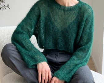 May include: A dark green, long-sleeved, loose-knit jumper with a fuzzy texture and a round neckline. The jumper is semi-sheer, revealing a dark V-neck garment underneath, and is paired with dark grey trousers featuring a visible waistband.