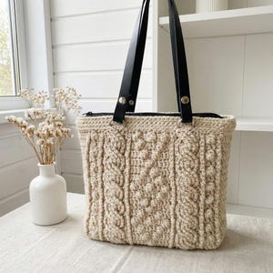 Puede incluir: Bolso tote beige tejido a ganchillo con un diseño de punto trenzado texturizado. El bolso tiene asas de cuero negro y una cremallera en la parte superior. El bolso está sobre una mesa junto a un jarrón blanco con flores secas.