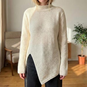 May include: Cream-colored knit sweater with a mock neck and long sleeves. The sweater features a unique asymmetrical hemline with a slit on the left side. The knitwear is a neutral color, suitable for various outfits.