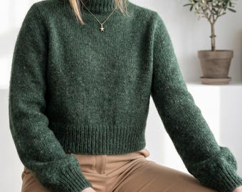 May include: A dark forest green long-sleeved jumper with a ribbed mock neck, cuffs, and hem. The knit fabric features a subtle heathered texture throughout. A delicate gold-toned necklace with a small star-shaped pendant rests against the neckline.