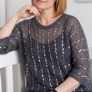 May include: A charcoal gray, sheer, loose-knit sweater featuring a round neckline and three-quarter length sleeves. The garment is embellished with vertical rows of small, reflective silver sequins that create a subtle shimmering effect throughout.
