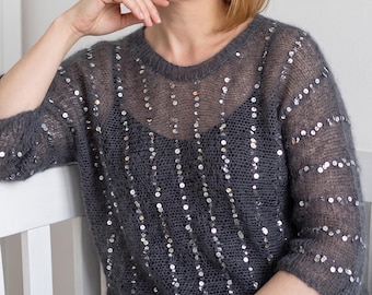 May include: A charcoal grey, sheer, loose-knit jumper featuring a round neckline and three-quarter length sleeves. The garment is embellished with vertical rows of small, reflective silver sequins that create a subtle shimmering effect throughout.