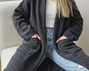 May include: A long, charcoal grey open-front cardigan featuring long sleeves and two large patch pockets on the front. The garment is worn over a white ribbed tank top and light blue denim jeans with a distressed knee detail.