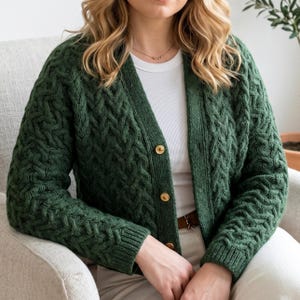 May include: A dark forest green cable knit cardigan featuring a V-neckline, long sleeves with ribbed cuffs, and a front button closure with three gold-toned circular buttons. The knit pattern consists of vertical braided textures throughout.