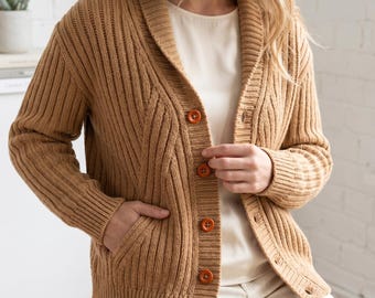 May include: A tan ribbed knit cardigan featuring a shawl collar, long sleeves, and five round brown buttons down the front. The garment includes two side pockets and is worn over a plain cream-coloured top with matching cream-coloured trousers.
