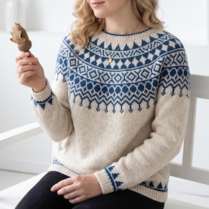 May include: A long-sleeved, crew-neck jumper in a light beige colour featuring a dark blue geometric pattern around the yoke and cuffs. The knit garment has a ribbed neckline, cuffs, and hem, showcasing a detailed, repeating motif of diamonds and stylized shapes across the chest and shoulders.