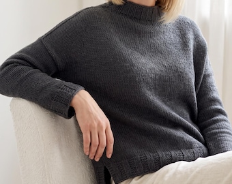 May include: A dark charcoal grey long-sleeve jumper featuring a ribbed mock neck collar, ribbed cuffs, and a ribbed hemline. The jumper has a relaxed fit with dropped shoulders and a small side slit at the bottom hem, crafted from a knit fabric.