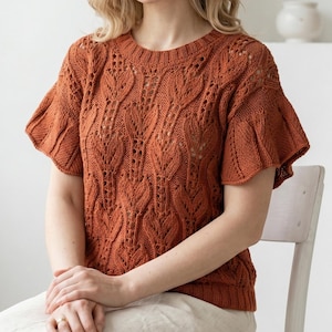 May include: A rust-colored knitted top with short, flared sleeves. The top features a detailed leaf pattern throughout the body and sleeves. The round neckline and the sleeves are ribbed. The person is sitting on a white chair.
