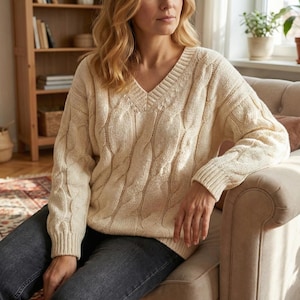 May include: Cream cable knit sweater with a V-neck. The sweater has a textured cable pattern and ribbed cuffs. The garment is made of a soft, warm material, suitable for cooler weather. A classic, versatile piece of knitwear.