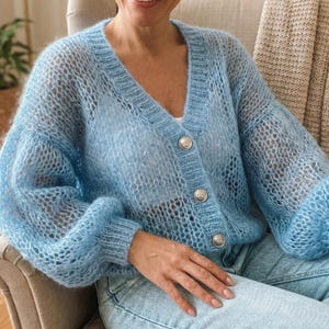 Może przedstawiać: Jasnoniebieski, ażurowy kardigan z dekoltem w serek i trzema srebrnymi guzikami. Kardigan ma długie, pękate rękawy i delikatny, przewiewny wygląd. Ubranie jest noszone z jasnoniebieskimi dżinsami.